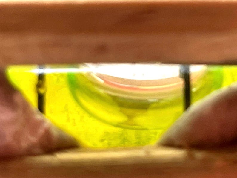 May include: A close-up view of a level tool, showing a yellow liquid bubble within a curved glass vial. The vial is set within a wooden frame, with the bubble centered, indicating a level surface.