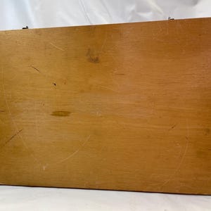 May include: A rectangular wooden box with a light brown finish. The box has visible scratches and wear, suggesting it has been used. The wood grain is visible. The box is closed and has a metal clasp.