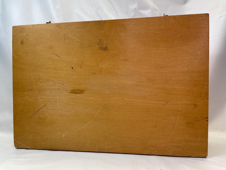 May include: A rectangular wooden box with a light brown finish. The box has visible scratches and wear, suggesting it has been used. The wood grain is visible. The box is closed and has a metal clasp.