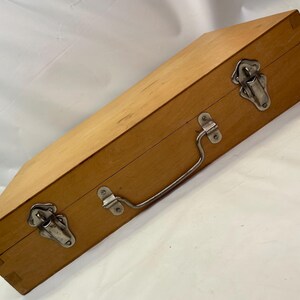 May include: A rectangular wooden box with a light brown finish. It features two metal latches, a metal handle, and metal hinges. The box is closed and appears to be empty. The box is likely used for storage or transport.