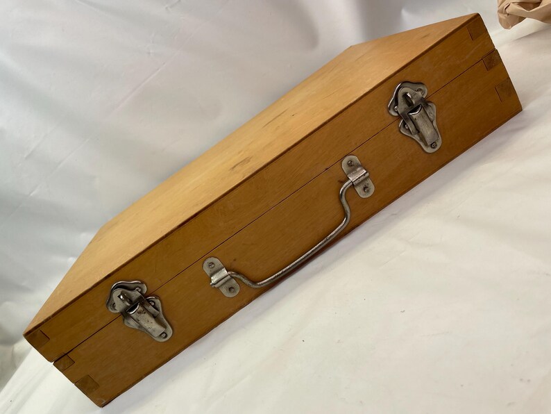 May include: A rectangular wooden box with a light brown finish. It features two metal latches, a metal handle, and metal hinges. The box is closed and appears to be empty. The box is likely used for storage or transport.