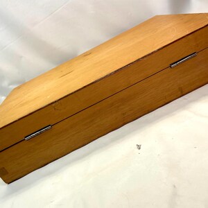 May include: A closed, rectangular wooden box with a light brown finish. The box has a hinged lid and metal hardware. The wood grain is visible, and the box appears to be well-constructed. The box is angled in the image.