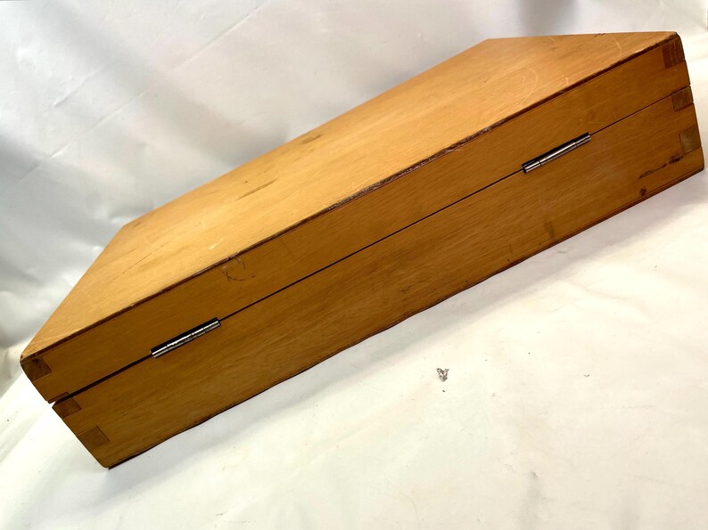 May include: A closed, rectangular wooden box with a light brown finish. The box has a hinged lid and metal hardware. The wood grain is visible, and the box appears to be well-constructed. The box is angled in the image.