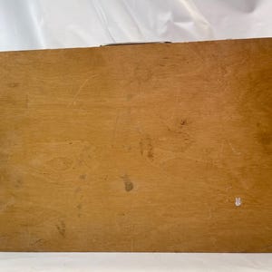May include: A rectangular wooden box with a light brown finish. The box has a metal handle on the top and shows signs of wear, including scratches and small dark spots. The wood grain is visible.