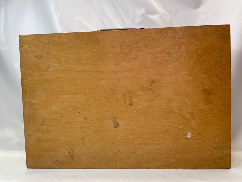 May include: A rectangular wooden box with a light brown finish. The box has a metal handle on the top and shows signs of wear, including scratches and small dark spots. The wood grain is visible.