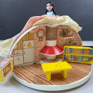 May include: A vintage dollhouse playset with a doll on top. The playset features a kitchen scene with a red and white stove, a yellow table, and a yellow shelf. The doll is wearing a blue and white dress.