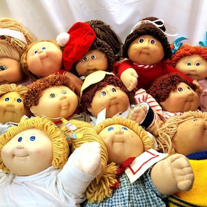 May include: A group of Cabbage Patch Kids dolls with different hairstyles and clothing. Some dolls are wearing hats, bows, and sweaters. The dolls have round faces, rosy cheeks, and blue eyes. The dolls are arranged closely together.