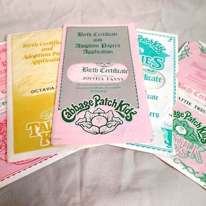May include: A collection of five Cabbage Patch Kids birth certificate and adoption papers application documents in pink, yellow, and white. Each paper features the Cabbage Patch Kids logo, text, and a cutout window displaying names like Joyvita Fanny.