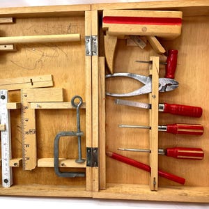 May include: A vintage wooden tool kit, open to reveal various carpentry tools. The kit includes a mallet, ruler, pliers, screwdrivers, and a clamp. The tools are arranged within the wooden box, showcasing a collection of essential instruments for woodworking and repair.
