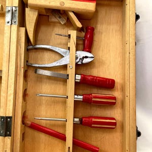 May include: Wooden toolbox with various hand tools. Includes a red-handled pliers, screwdrivers, and a pencil. The box is open, revealing the tools neatly arranged inside. The wood has a natural finish.