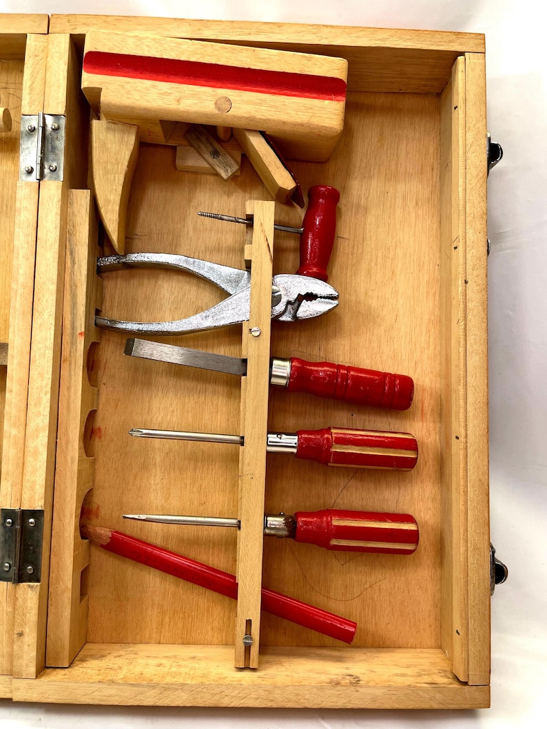 May include: Wooden toolbox with various hand tools. Includes a red-handled pliers, screwdrivers, and a pencil. The box is open, revealing the tools neatly arranged inside. The wood has a natural finish.