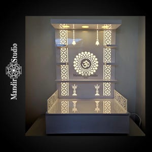 May include: A white, illuminated Mandir Studio shrine with intricate cut-out designs. The shrine features shelves, decorative lamps, and a central Om symbol. The design includes hanging bells and a decorative base, creating a serene and elegant aesthetic.