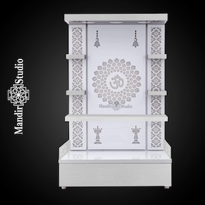 May include: A white wooden temple with intricate floral and Om symbol carvings. The temple features shelves, decorative bells, and the text "Mandir Studio". The design is symmetrical and elegant, suitable for home decor.