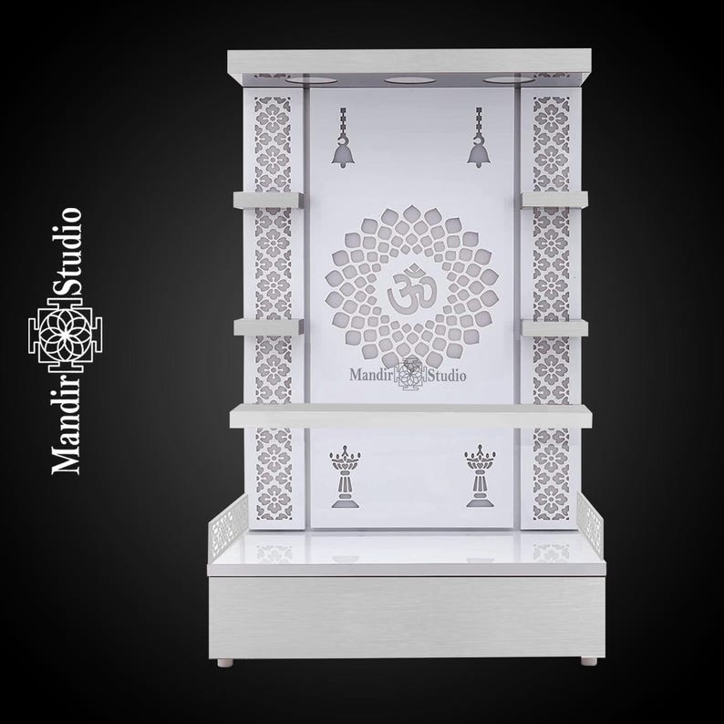 May include: A white wooden temple with intricate floral and Om symbol carvings. The temple features shelves, decorative bells, and the text "Mandir Studio". The design is symmetrical and elegant, suitable for home decor.