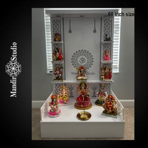 May include: A white wooden altar with multiple shelves, adorned with colorful Hindu deity statues. The altar is 60 inches in size and features intricate cut-out designs and the text "Mandir Studio".