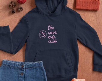 May include: Navy blue hoodie with the text "the cool kids club" in pink, a denim skirt, orange knit beanie, and fox-themed socks. An apple and pinecones are also present. The items are arranged on a wooden surface.