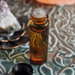 May include: Amber glass essential oil bottle with a black cap. The bottle is filled with a golden liquid. The cap is off, revealing the opening. The bottle is on a decorative surface with a blurred background.