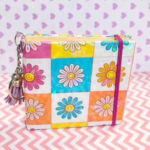 Könnte beinhalten: Eine bunte, quadratische Geldbörse mit einem Gänseblümchenmuster. Die Geldbörse hat ein Raster aus rosa, gelben, blauen und orangen Quadraten, jedes mit einem Gänseblümchen-Design. Ein lila Gummiband und ein Schlüsselanhänger mit Quaste sind befestigt.