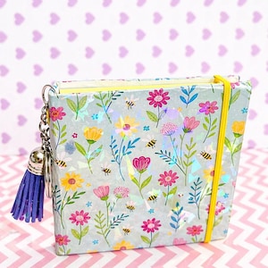 May include: A notebook with a floral design and a blue tassel keyring. The notebook has a light blue cover adorned with flowers, bees, and holographic details. A yellow elastic band holds it closed.