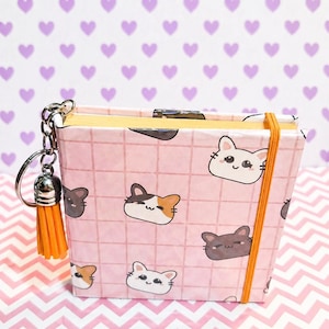 May include: A small, pink notebook with a grid pattern and repeating cat face illustrations. The notebook has an orange elastic band and a matching tassel keychain. The background features a repeating heart pattern.