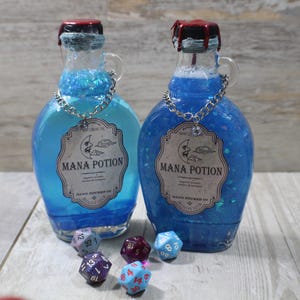 Może przedstawiać: Dwie szklane butelki wypełnione niebieskim płynem z napisem "Mana Potion", ze srebrnym łańcuszkiem i ozdobną etykietą. Butelki mają czerwone i czarne korki. Kilka wielościennych kości jest rozrzuconych przed butelkami.