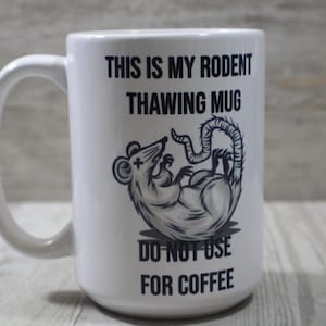 May include: White ceramic mug with a black graphic of a rat and the text "THIS IS MY RODENT THAWING MUG DO NOT USE FOR COFFEE". The mug has a large handle and is suitable for hot beverages.
