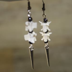 May include: A pair of dangle earrings featuring white animal vertebrae bones alternating with small, round, polished black beads. Each earring is finished with a silver-toned metal spike charm at the bottom and hangs from a black metal hook.