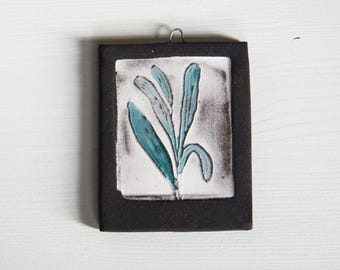 May include: A small, square ceramic tile with a black frame and a white background. A blue and green stylized plant is painted on the tile.
