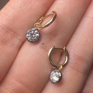 May include: Two gold hoop earrings, each featuring a round, diamond-like stone pendant set in a dark metal bezel. The earrings are shown against a neutral backdrop. The brand name "SOLITAIRE DIAMONDS" is visible.