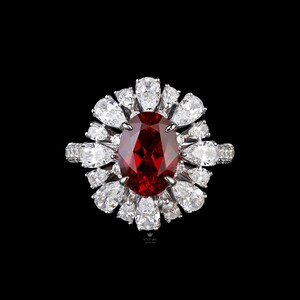 May include: A dazzling ring featuring a large, oval-cut, deep red gemstone as the focal point, surrounded by a halo of pear-shaped and round white gemstones. The ring's band is also adorned with smaller white gemstones, set against a black background.