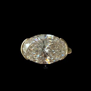 May include: A ring featuring a large, oval-cut, clear faceted gemstone set in a gold-toned metal band. The stone displays multiple internal reflections and light refractions. Below the ring, the text SOLITAIRE DIAMOND is printed in small letters.