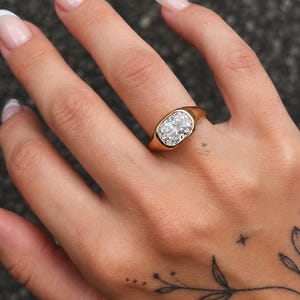 Op de afbeelding: Een gouden ring met een grote, heldere edelsteen in kussenslijpsel, gezet in een gladde, gepolijste zetting. De ringband is van massief goud met een afgerond profiel en het metalen oppervlak reflecteert het licht, wat de helderheid benadrukt.