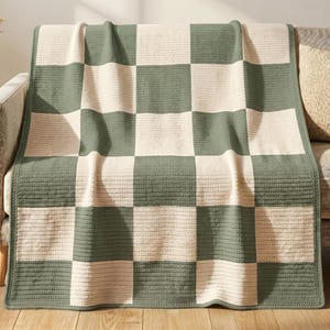May include: A rectangular throw blanket featuring a large-scale checkered pattern in alternating sage green and cream colors. The blanket has a visible ribbed or knit texture throughout the entire surface, creating a consistent grid design.