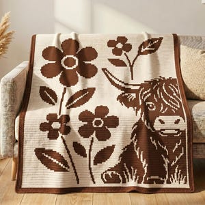 May include: A cream-colored woven throw blanket featuring a dark brown pixelated pattern of a Highland cow and several flowers. The blanket has a solid dark brown border and a textured knit construction, displaying a repeating geometric design.