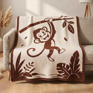 May include: A cream-coloured crochet blanket featuring a large brown graphic of a monkey hanging from a branch. The design includes brown tropical leaves at the bottom corners and a solid brown border framing the entire rectangular textile piece.
