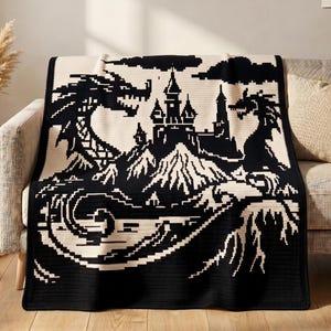 May include: A black and cream-colored throw blanket featuring a pixelated graphic design of a central castle surrounded by mountains, two large dragons on either side, and stylized waves at the bottom, all rendered in a high-contrast knit pattern.