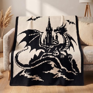 May include: A rectangular knit throw blanket featuring a high-contrast black and cream pixelated design. The graphic depicts a large dragon perched on a rocky cliff in front of a tall, pointed castle tower, with two small bats flying in the sky.