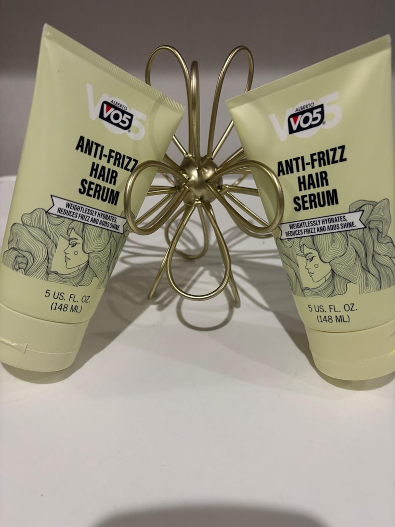 May include: Two tubes of Alberto VO5 Anti-Frizz Hair Serum, each containing 148 ml, are displayed. The tubes are light yellow with black text and a drawing of a woman with wavy hair. They are held by a gold-coloured, decorative metal holder.
