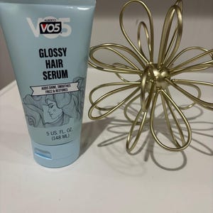 May include: A light blue tube of Alberto VO5 Glossy Hair Serum. The tube has black text and a drawing of a woman's face. The tube is 5 US fl oz (148 mL). A gold metal flower-shaped decoration is next to the tube.