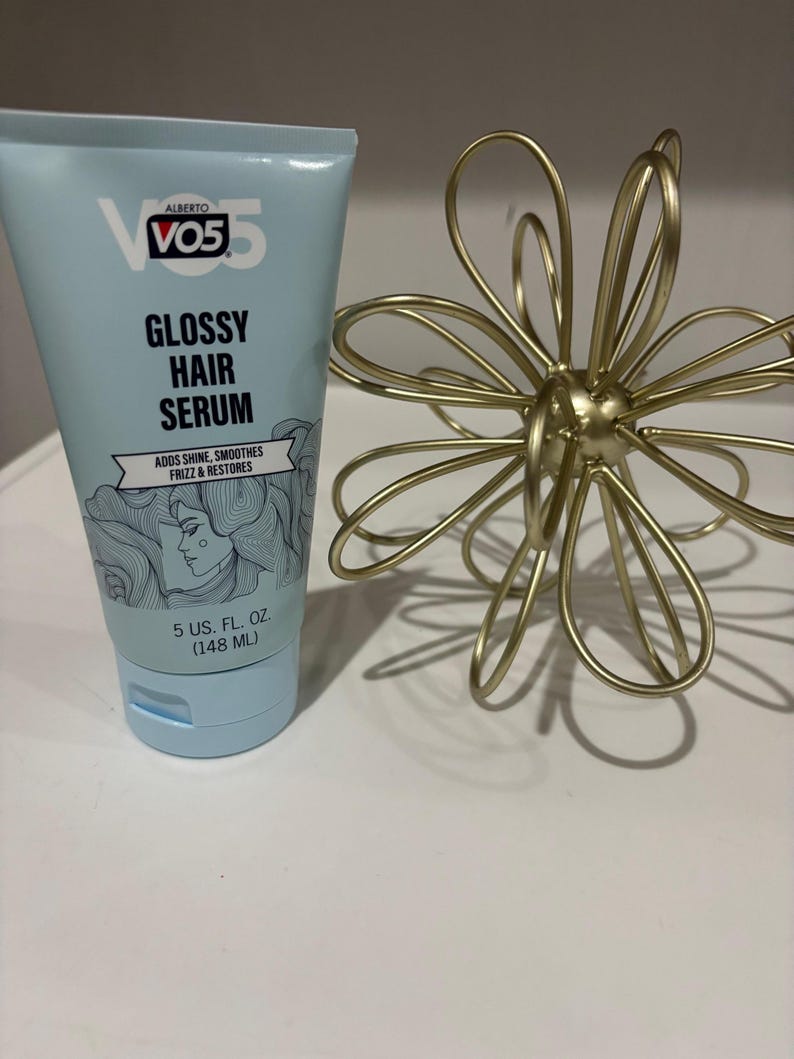 May include: A light blue tube of Alberto VO5 Glossy Hair Serum. The tube has black text and a drawing of a woman's face. The tube is 5 US fl oz (148 mL). A gold metal flower-shaped decoration is next to the tube.