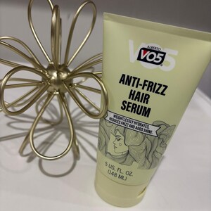 May include: A light yellow tube of VO5 Anti-Frizz Hair Serum, with text and a woman's face illustration. The tube is 148 ml. A gold-coloured, wire floral sculpture is to the left of the tube.