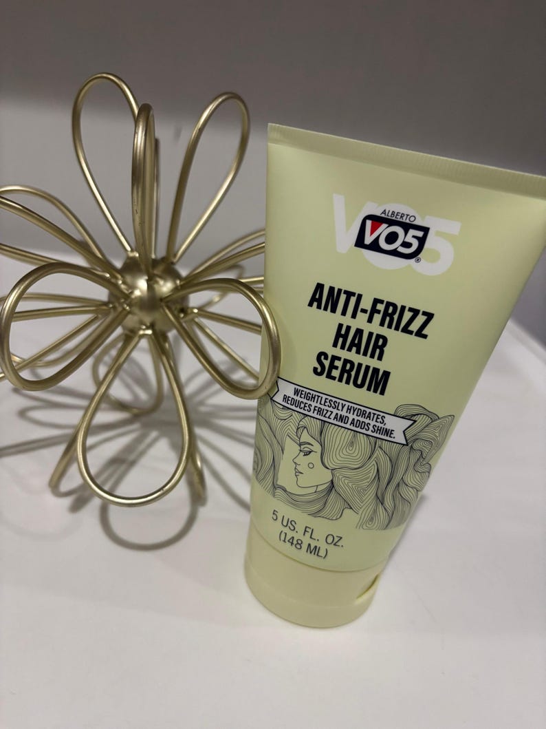 May include: A light yellow tube of VO5 Anti-Frizz Hair Serum, with text and a woman's face illustration. The tube is 148 ml. A gold-coloured, wire floral sculpture is to the left of the tube.