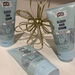 May include: Three light blue tubes of VO5 Glossy Hair Serum, each with a white cap and the product name in black. The tubes are arranged with a gold-colored, abstract flower-shaped decoration.