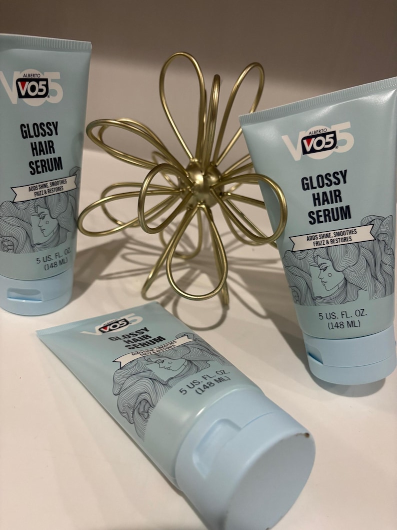 May include: Three light blue tubes of VO5 Glossy Hair Serum, each with a white cap and the product name in black. The tubes are arranged with a gold-colored, abstract flower-shaped decoration.