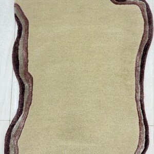 May include: A beige, irregularly shaped rug with a layered border in shades of brown and mauve. The rug has a soft, plush texture and is designed to add a decorative element to any room.