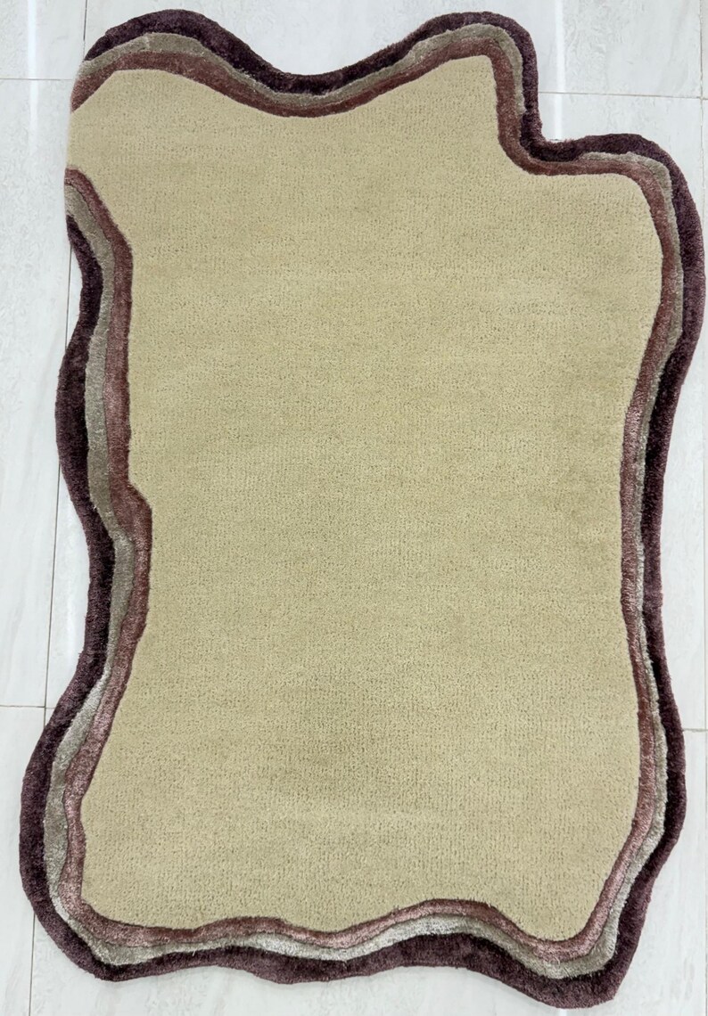 May include: A beige, irregularly shaped rug with a layered border in shades of brown and mauve. The rug has a soft, plush texture and is designed to add a decorative element to any room.