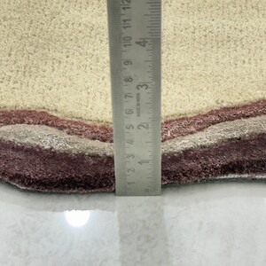 May include: Close-up of a beige and burgundy area rug with a wavy design. The rug has a textured surface and a layered pattern in shades of beige, gray, and burgundy. A ruler is placed vertically on the rug, showing measurements in inches.