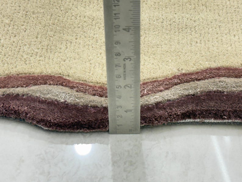 May include: Close-up of a beige and burgundy area rug with a wavy design. The rug has a textured surface and a layered pattern in shades of beige, gray, and burgundy. A ruler is placed vertically on the rug, showing measurements in inches.