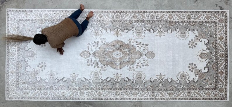May include: A large rectangular area rug featuring a central medallion and intricate floral border patterns in shades of beige, cream, and soft brown. The rug displays a symmetrical design with detailed motifs throughout the field and edges.