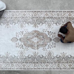 May include: A large rectangular area rug featuring a central medallion and intricate floral border patterns in shades of beige, cream, and soft gray. The design includes symmetrical motifs and detailed scrollwork across the entire surface.